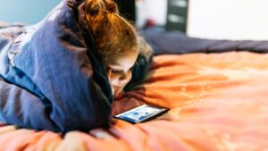 Criança assistindo celular na cama apoiada sobre os cotovelos